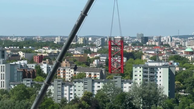 High-angle view of a mobile telescopic crane lifting a red lattice tower crane section against a sprawling, sunlit city skyline