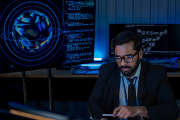 Fototapeta premium South Asian male developer in a white shirt uses a tablet device while surrounded by screens displaying algorithms in a dark tech office.