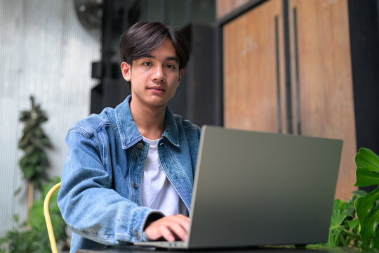 Young Asian non-binary man using laptop looking at camera wide
