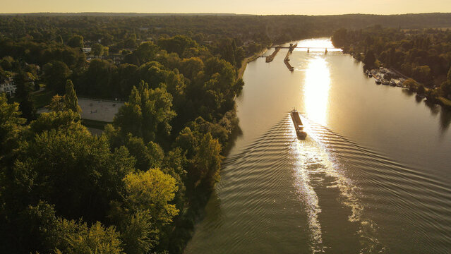 Aerial view of a barge navigating the Seine river during golden hour, surrounded by lush green forests and trees in Chartrettes, Ile-de-France, France.