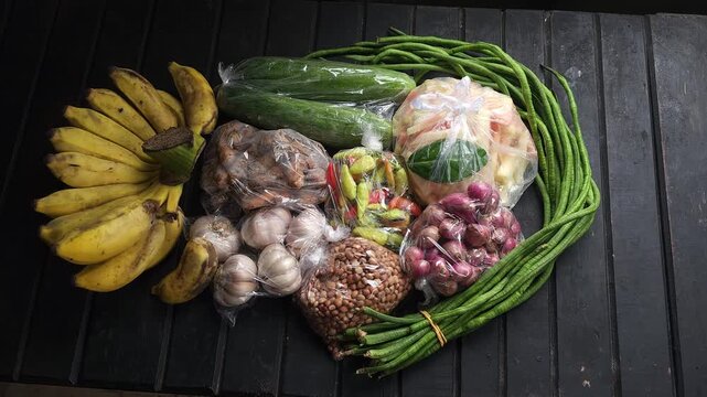 A collection of fresh produce including bananas, cucumbers, yardlong beans, garlic, peanuts, chilies, and shallots neatly arranged on a dark wooden table