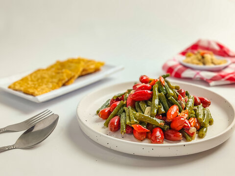 A dish of Fresh long beans and melinjo skin are served on a white plate with a minimalist table setting. A healthy, home-cooked vegetable dish with bright colors and a selective focus.