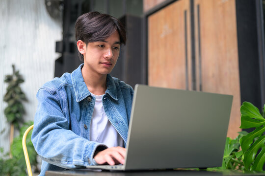 Young Asian non-binary man using laptop looking down at screen wide