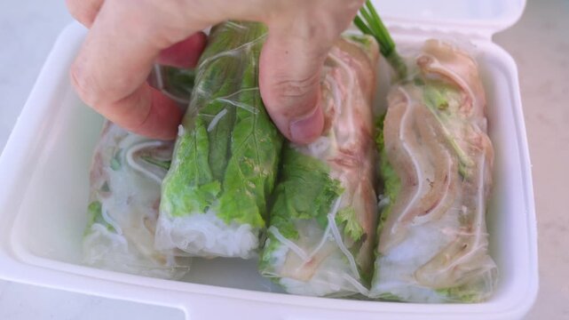 Close up of a person's hand taking a traditional vietnamese fresh spring roll, filled with pork, vermicelli, and herbs, from a white styrofoam take out container on a plain surface