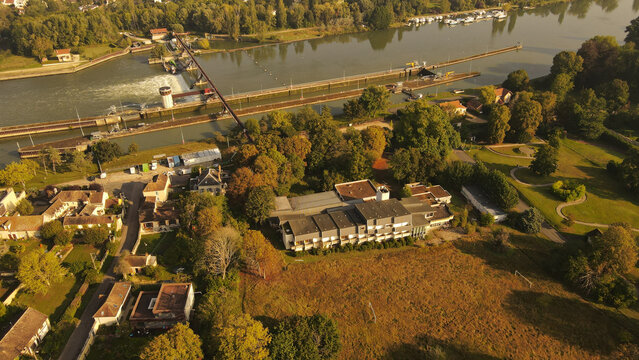 Aerial view of the Seine river lock system and surrounding buildings in the village of Chartrettes, featuring lush trees and golden hour light in Chartrettes, Ile-de-France, France.