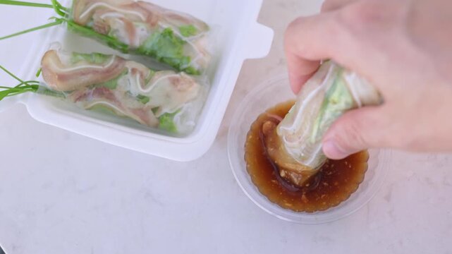 Human hand dipping a traditional vietnamese spring roll, filled with pork ear and fresh vegetables wrapped in rice paper, into a savory peanut dipping sauce from a takeout container