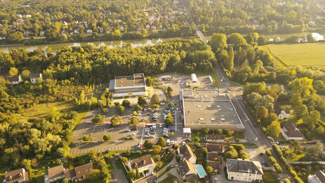 Aerial view of a commercial complex with parking lots, surrounded by lush green trees and residential houses near the Seine river in Chartrettes, Ile-de-France, France.