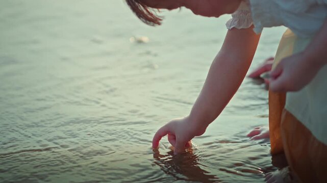 Close up of little Asian girl hand picking up small seashell from sea water on beach, cute curious child collecting sea shells during summer family vacation at sunset for travel, childhood.