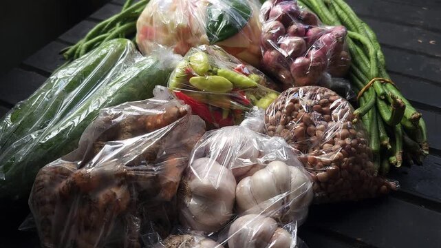 Assortment of fresh raw vegetables and spices like cucumbers long beans shallots garlic and chilies in plastic bags resting on a dark surface for food preparation