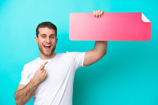 Young caucasian man isolated on blue background holding an empty placard and pointing it