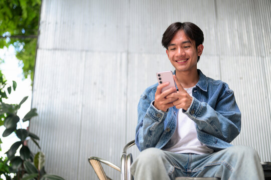 Young Asian non-binary man smiling using phone sitting outdoor wide