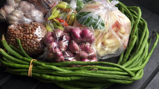 Fresh raw vegetables and cooking ingredients including yardlong beans shallots garlic peanuts and chilies wrapped in plastic bags on a wooden table
