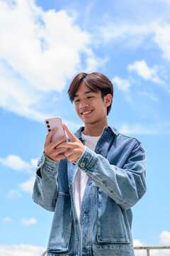 Young Asian non-binary man smiling using phone under blue sky portrait