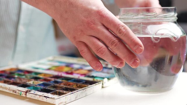 Closeup of artist hand placing water jar on desk beside watercolor paints