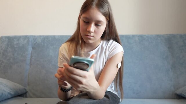 Teenage girl looking at her smartphone screen to monitor glucose levels using a health app. A white sensor patch is visible on her upper arm. Diabetes management, health technology, and telemedicine