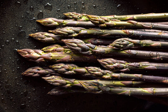 Purple asparagus sprinkled with water on a black background, focus on asparagus on the middle, close up view