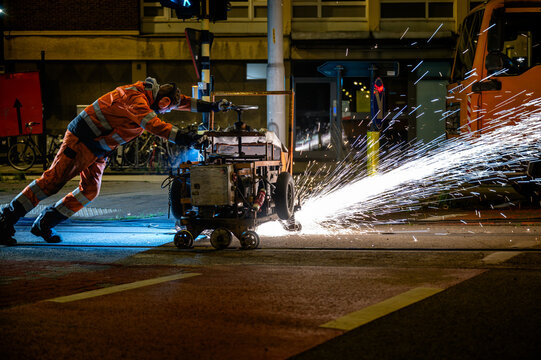 A worker grinds a tram rail at night with sparks flying across a city street beside an orange truck, illustrating night-time railway track maintenance and road works.