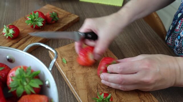 Slicing fresh organic strawberries on olive wood cutting board in kitchen. Hands of chef cutting summer berries with knife next to enamel colander reflecting culinary creativity and food styling