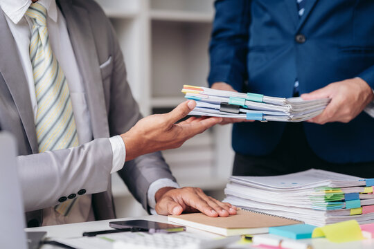 Two men are standing in front of a desk with a pile of papers