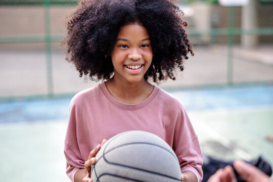 Smiling girl with curly hair holds basketball on outdoor court.
