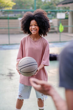 Smiling girl with curly hair holds basketball on outdoor court.