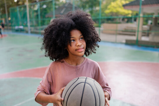 Girl with basketball looks up on outdoor court.