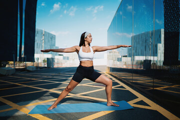 Obraz premium Confident woman holding warrior pose, centered on mat, arms fully extended, aligned with city symmetry, showing mental strength, body tone, and flow of mindful digital movement.