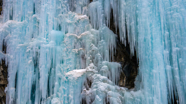 Aerial view of a massive frozen waterfall with intricate blue icicles hanging from a rugged rock face in Khunjerab National Park, Gilgit, Pakistan.