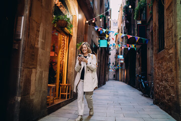 Obraz premium Full length smiling woman using smartphone walking on street. Mature traveler checking booking, mobile banking, online service, digital nomad lifestyle, authentic representation, copy space