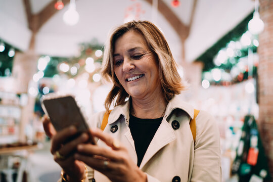 Middle aged woman interacting with smartphone touchscreen in cozy interior, happy female user browsing mobile content, authentic portrait of connectivity and positive emotions.