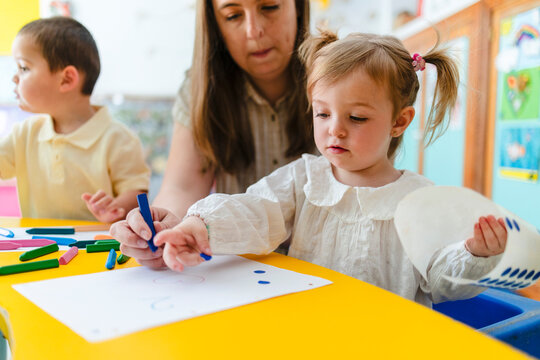 Teacher assisting young child with crayon drawing in kindergarten