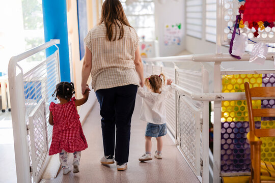 Teacher guiding multiracial toddlers walking in daycare hallway