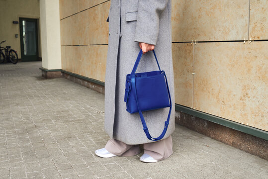 Person stands on sidewalk with gray coat and blue bag in urban area 
