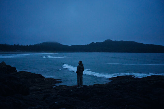 Person stands on rocks by ocean at dusk observing the waves