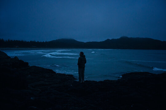 Person stands on rocks by ocean at dusk observing the waves