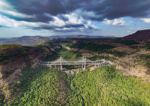 Aerial view of the Mumbai-Pune Expressway Missing Link Project cable-stayed bridge under construction, spanning a deep forested valley with dramatic clouds in Khopoli, Maharashtra, India.