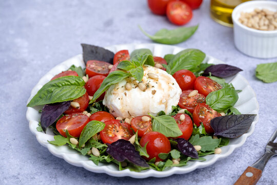 Fresh burrata salad served with ripe cherry tomatoes, mixed greens, fresh basil leaves, and toasted pine nuts on a gray background. Healthy eating concepts, and Italian cuisine. side view