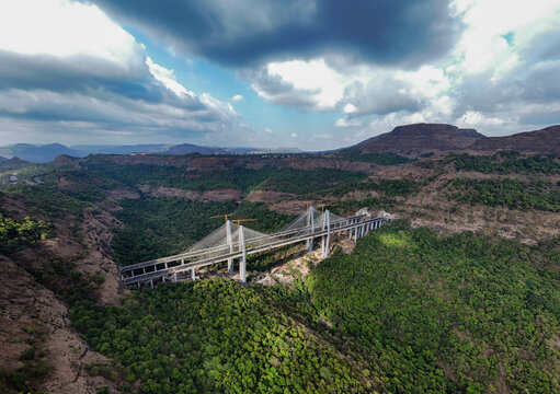 Aerial view of the Mumbai-Pune Expressway Missing Link Project bridge spanning a lush green valley with cranes and hills under a cloudy sky in Khopoli, Maharashtra, India.