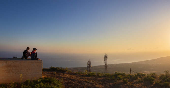 Mother and son sitting on concrete platform at sunrise, looking at scenic panoramic view towards Malia town from Mount Edheri in Crete, Greece. Family hiking and travel concept