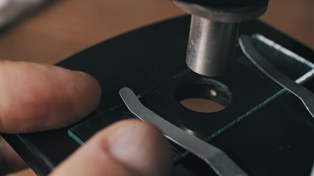 Hands place a glass slide on a microscope stage and add a drop of liquid using a pipette. This represents meticulous scientific research and analysis in a laboratory setting.