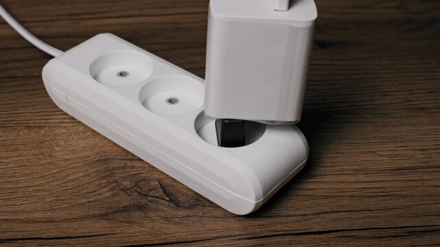 A hand plugs a large white power adapter with a black Europlug base into a white triple socket extension cord. The setup rests on a dark wood grain desk connecting equipment to power.