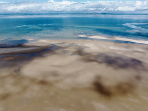 Aerial view of the Moray Firth Coast, featuring expansive sandy tidal flats, shallow turquoise waters, and dramatic cloud shadows cast across the beach in Nairn, Scotland, United Kingdom.
