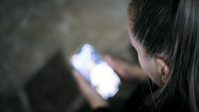 Over-the-shoulder view of a woman holding a smartphone with a bright screen glowing in a dark room. The anonymous female browses the internet with her face unseen.