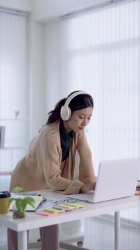Young Asian woman wearing headphones working on laptop at home office desk with organized workspace