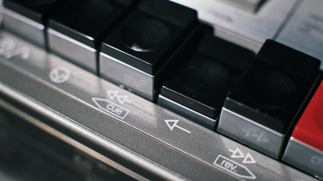 Angled close-up of a finger pushing a black square play control on an analog audio deck. The brushed metal surface shows printed icons and transport keys in use.