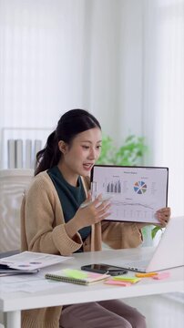 Young Asian businesswoman presenting finance report charts during video call in modern bright home office workspace