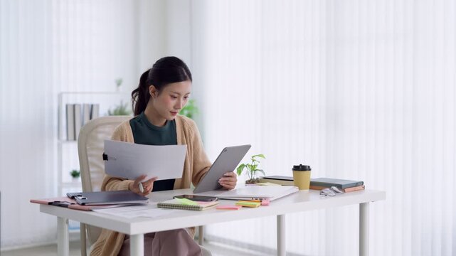 Young Asian professional woman comparing paper documents and digital tablet while working at bright home office desk
