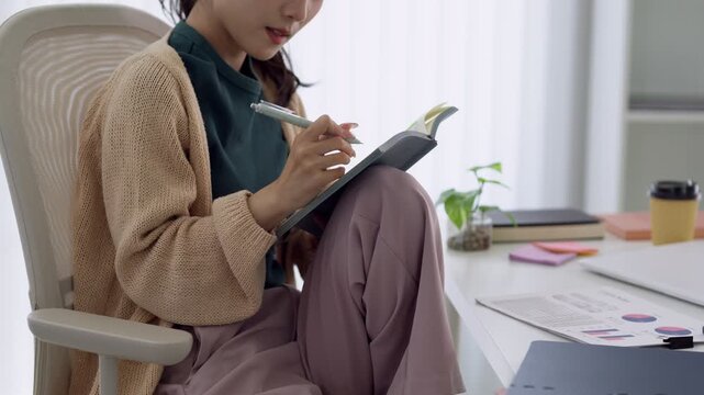Young Asian professional writing notes in planner while reviewing business charts at bright home office desk