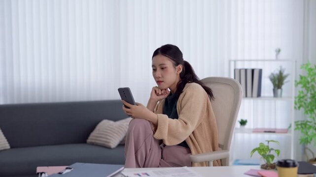 Young Asian woman looking at smartphone with concerned expression while working remotely in bright modern home office