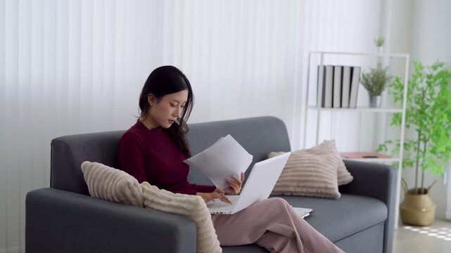 Young Asian professional reviewing documents and using laptop while working remotely from home on living room sofa